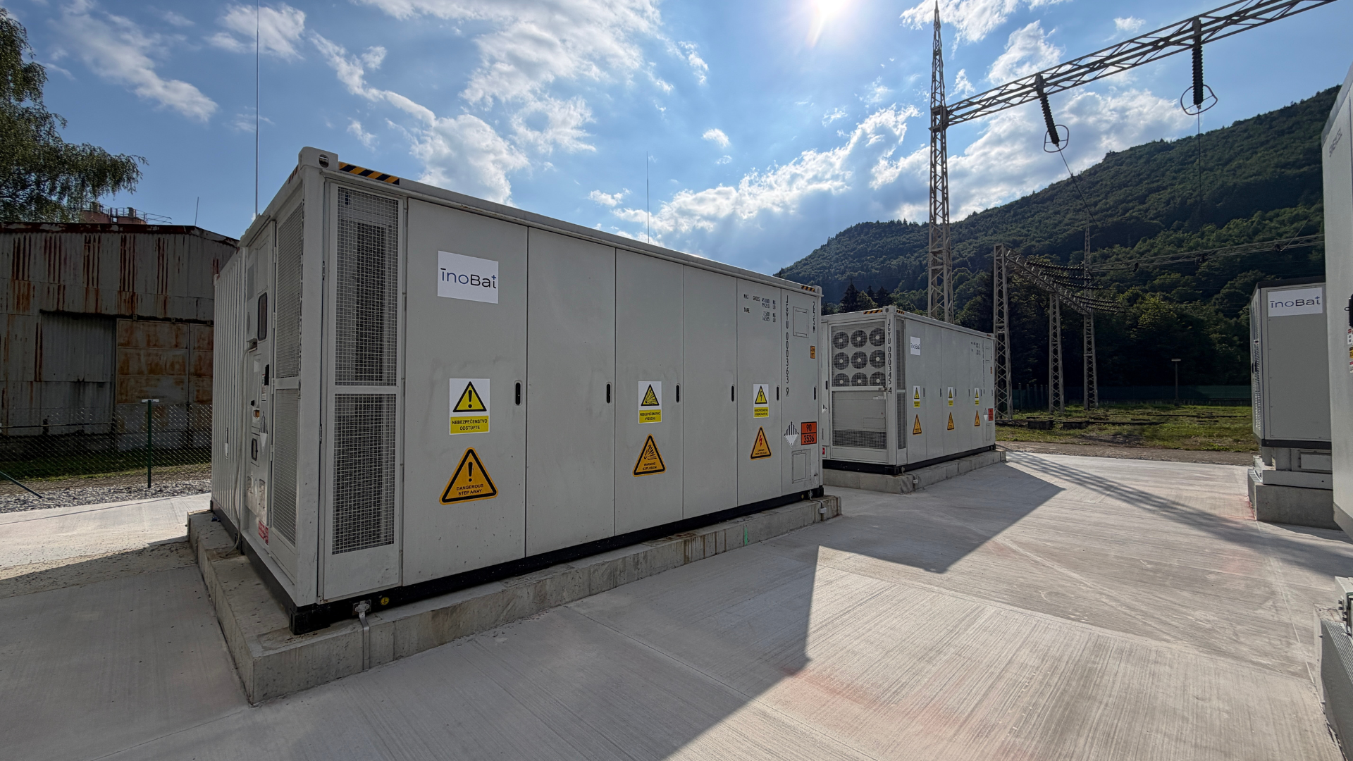 Containerised BESS units on concrete foundations at OFZ Oravský Podzámok, with the substation and forested hills behind